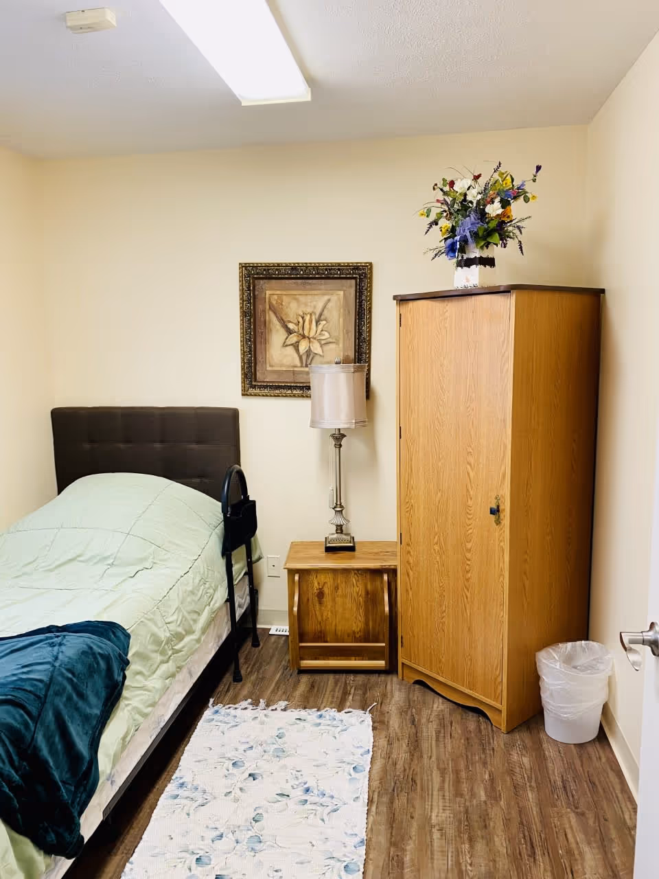 Small furnished bedroom with a single bed, nightstand and lamp, a wooden wardrobe topped with flowers, and a decorative rug on wood flooring.