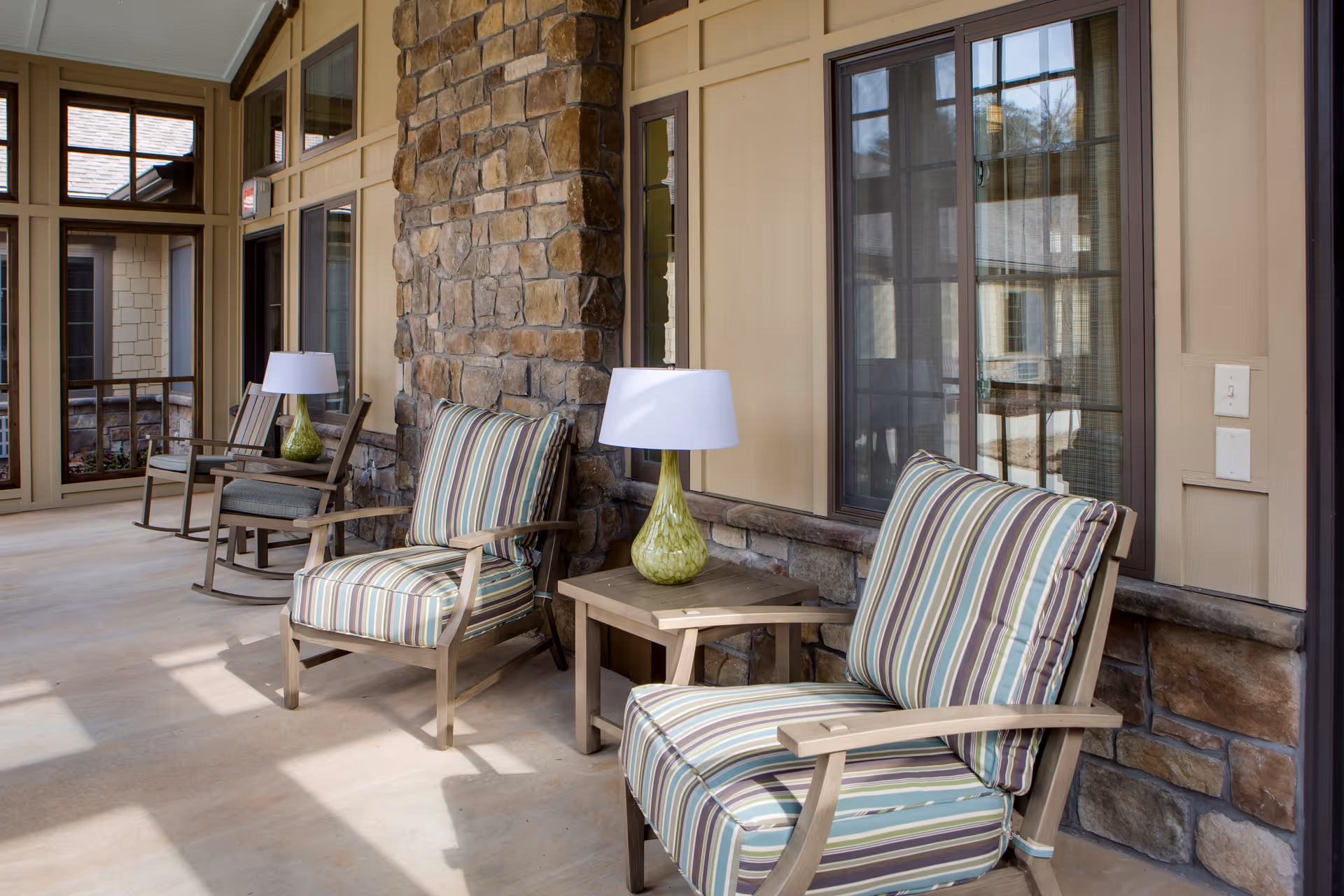 A covered outdoor patio area with cushioned wooden chairs featuring striped upholstery, a small wooden side table with a green glass lamp, and a stone wall with windows and beige siding.