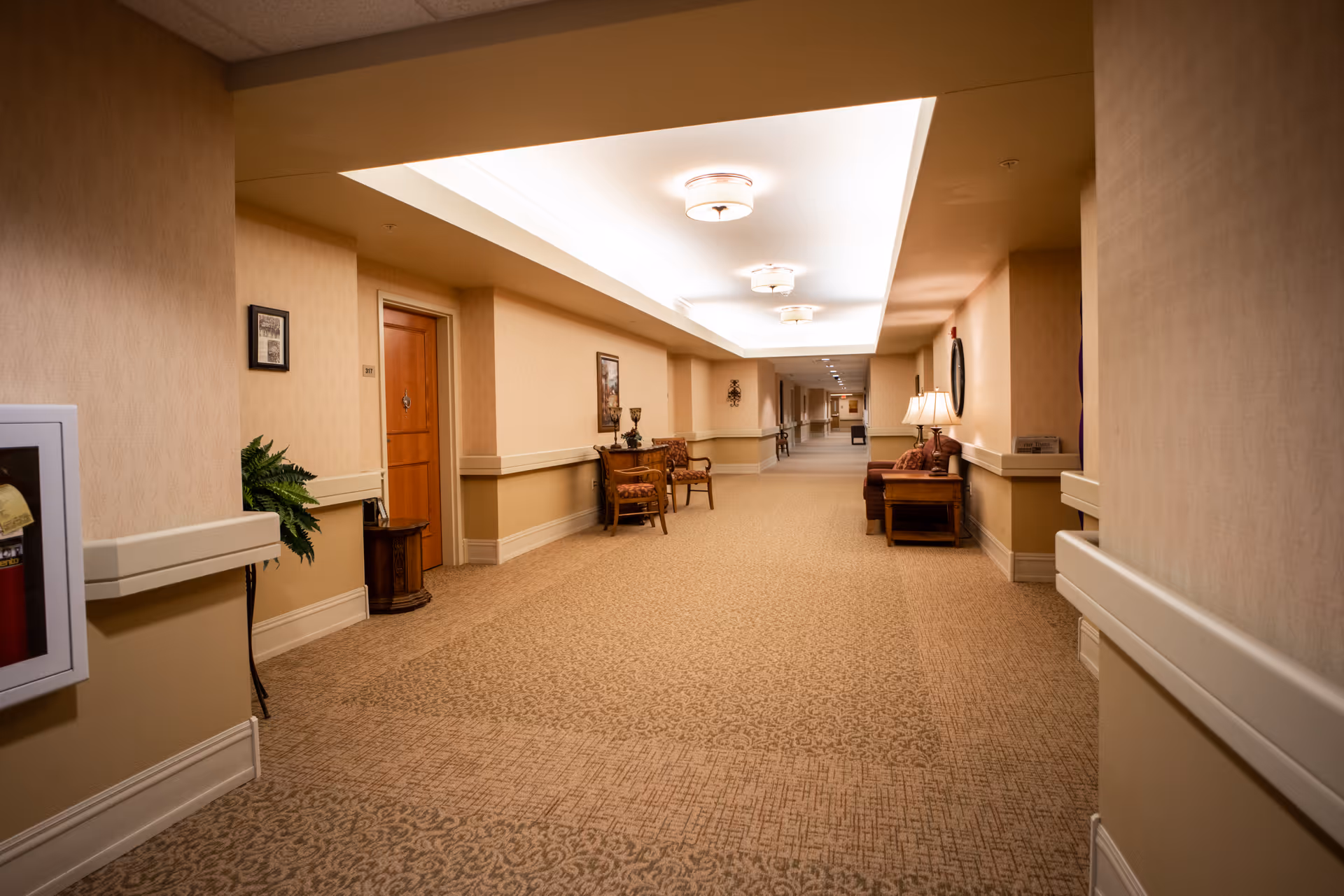 A well-lit, carpeted interior hallway with seating areas, side tables, framed art, and multiple apartment doors.