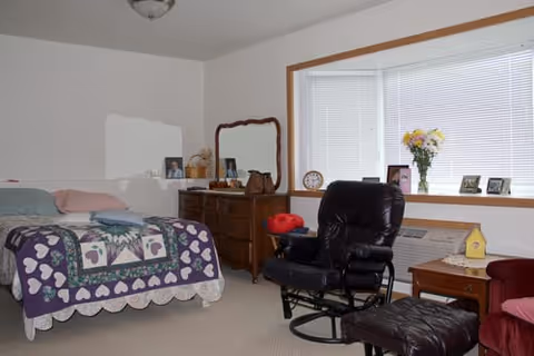 A furnished bedroom with a bed covered in a heart-patterned quilt, wooden dresser, black leather recliner and a bay window with blinds and flowers.