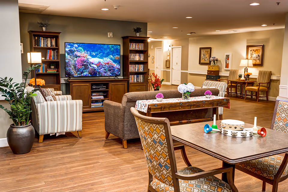 A cozy senior living facility common area with a large flat-screen TV displaying an underwater coral scene, flanked by two wooden bookshelves filled with books and DVDs. The room features comfortable seating including a striped armchair, a brown sofa, and patterned chairs around a wooden table with musical instruments on it. The space is decorated with plants, flowers, framed artwork, and warm lighting.