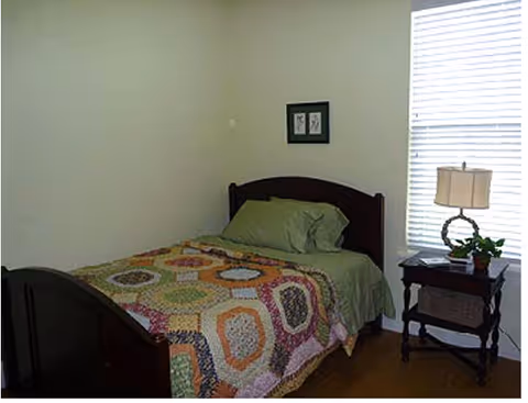 A bedroom with a single bed, patchwork quilt, green pillows, nightstand, lamp, and blinds.