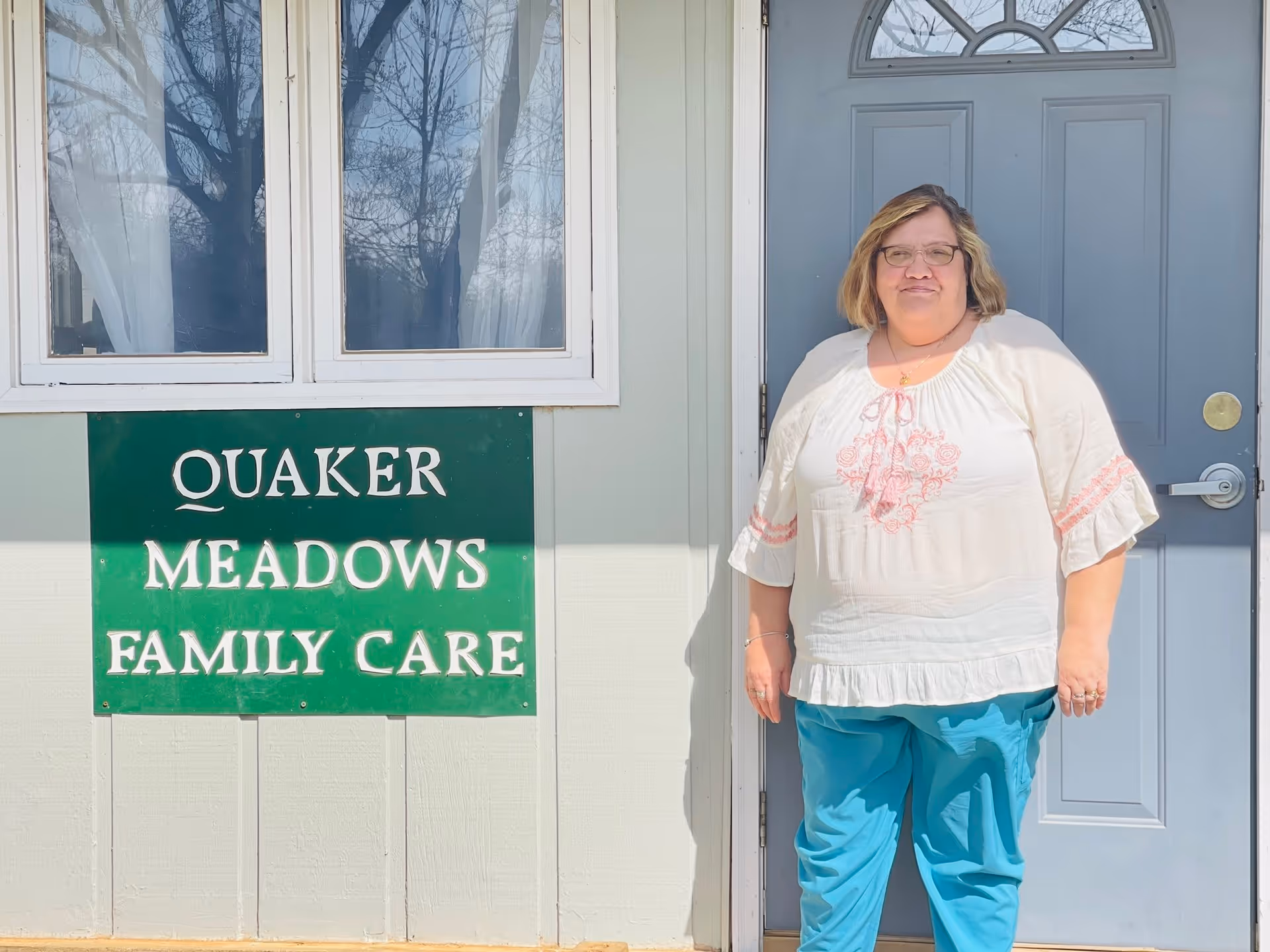 A woman with glasses wearing a white blouse with pink embroidery and blue pants stands in front of a gray door next to a green sign that reads 'Quaker Meadows Family Care'. The building exterior is light-colored with two windows above the sign.