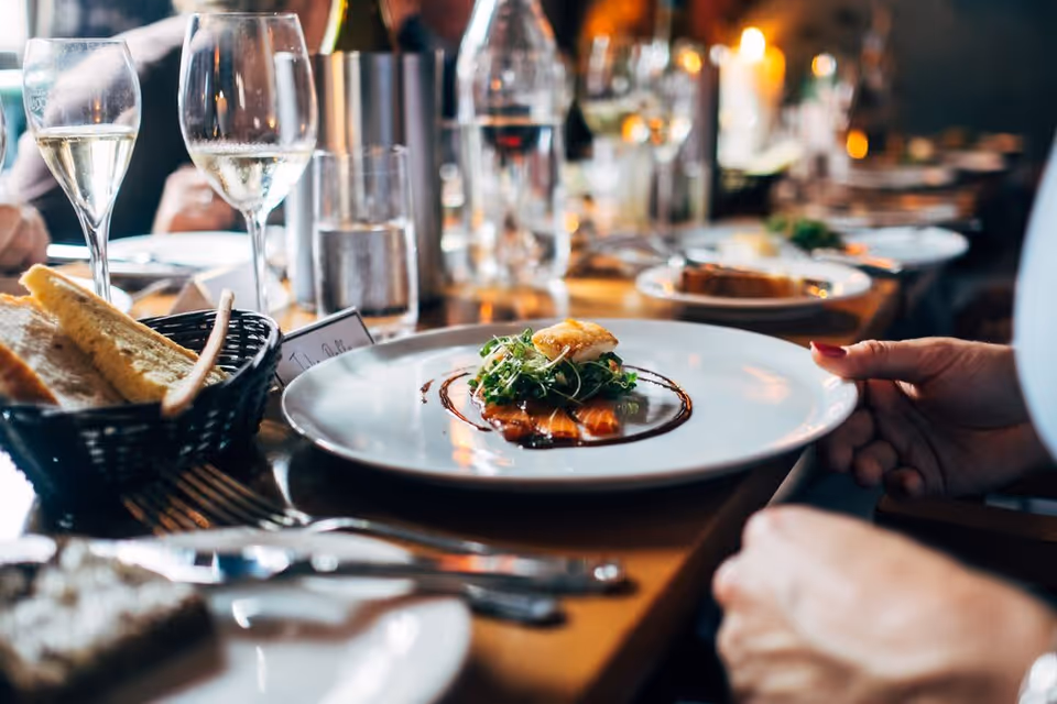 A close-up view of a dining table set with plates of gourmet food, glasses of wine, a basket of bread, and cutlery. A person's hand is visible holding a plate with a small, elegantly plated dish. The background shows more plates and glasses on the table in a warmly lit dining setting.