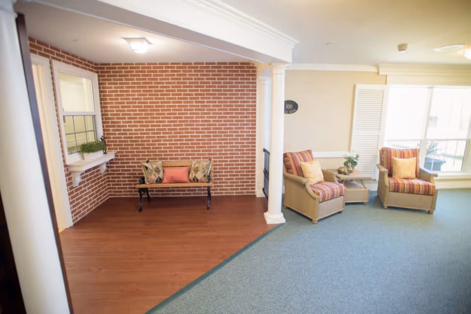 A cozy indoor seating area with a small bench against a red brick wall on the left and two cushioned armchairs with striped upholstery and yellow pillows on the right. A small table with a flower arrangement is placed between the armchairs. The floor transitions from wood to carpet, and there is a large window letting in natural light.