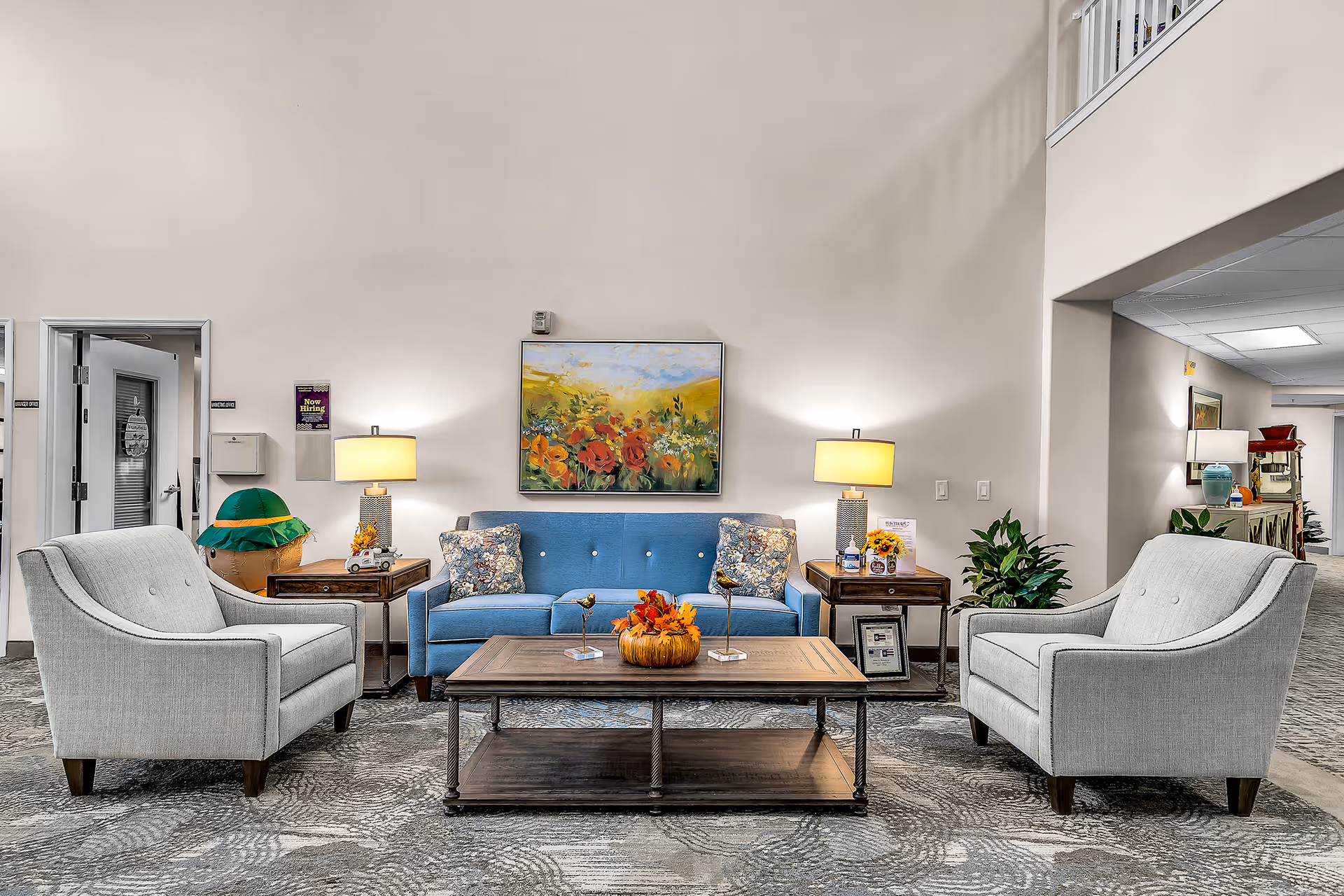 A cozy seating area in a senior living facility featuring a blue sofa with floral cushions, two light gray armchairs, a wooden coffee table decorated with a fall-themed centerpiece, two side tables with lamps, a colorful floral painting on the wall, and a carpeted floor with a patterned design.