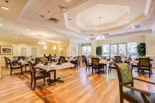 A spacious dining room with multiple tables and chairs arranged neatly. The room features large windows allowing natural light to fill the space, elegant ceiling design with recessed lighting and chandeliers, and framed artwork on the walls. The floor is wooden, and the overall atmosphere is bright and welcoming.