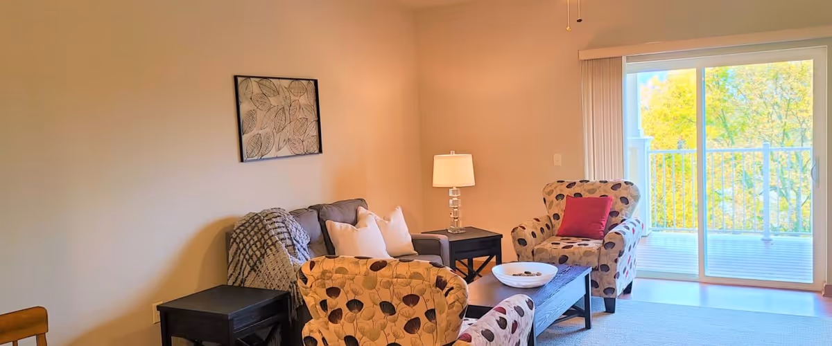 A cozy living room with a gray sofa adorned with white pillows and a patterned throw blanket, two matching patterned armchairs with a red cushion on one, a dark wooden coffee table with a decorative bowl, a side table with a lamp, and a large sliding glass door leading to a balcony with a view of green trees.