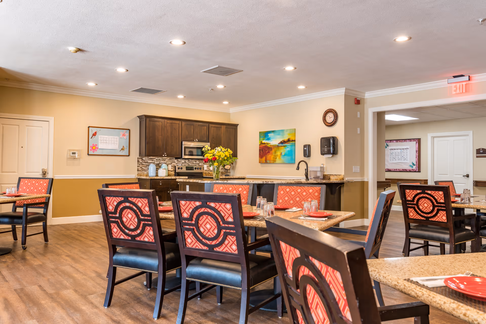A bright dining room in McMinnville Memory Care featuring multiple tables with red patterned chairs. The tables are set with red plates and glasses. In the background, there is a kitchen area with dark wood cabinets, a stove, microwave, and a countertop with a vase of yellow flowers. The walls are painted beige with white trim, and there are colorful paintings and a clock on the walls. The floor is wood, and ceiling lights illuminate the space.