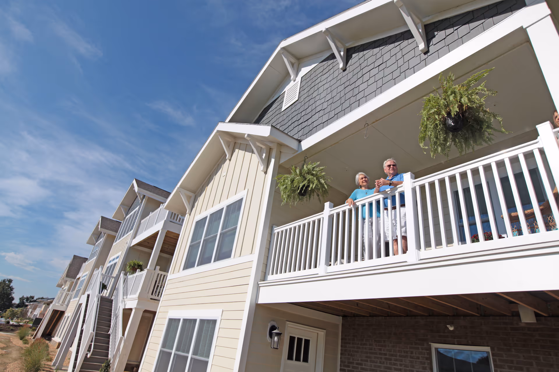An elderly couple standing on the balcony of a multi-story residential building with beige siding and white railings, under a blue sky with some clouds. Two hanging fern plants are visible on the balcony.