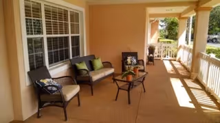 Covered outdoor patio area with wicker seating including a loveseat and two chairs, a glass-top coffee table with potted plants, and a railing along the edge. The patio is attached to a building with peach-colored walls and large windows, and there is greenery and a parked car visible in the background.