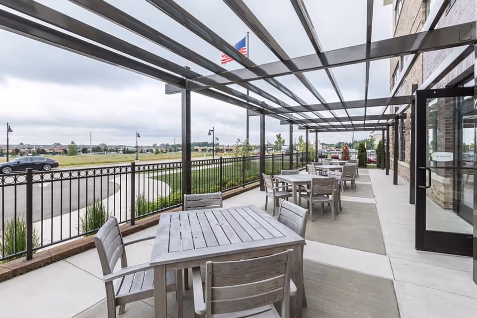 Outdoor patio area with multiple wooden tables and chairs under a transparent pergola attached to a brick building. An American flag is visible in the background along with a parking lot and grassy area.