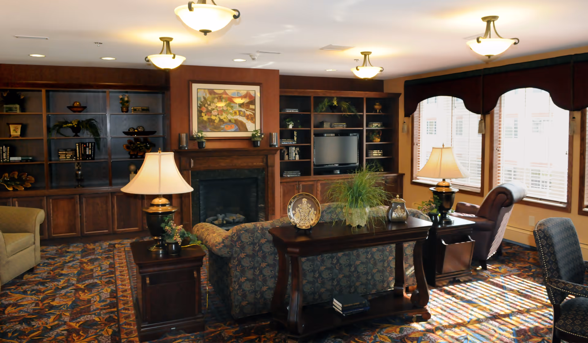 A cozy lounge with upholstered seating, table lamps, built-in bookshelves, a fireplace, and large windows.
