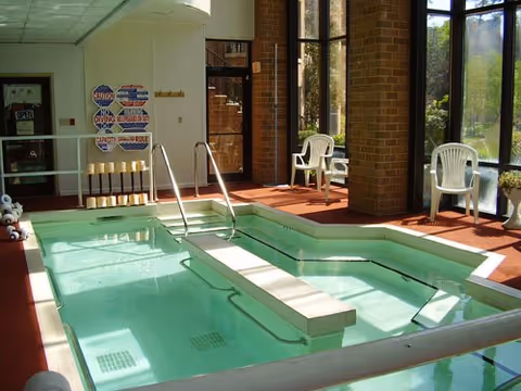 Indoor therapy pool with handrails, chairs, and large windows letting in sunlight.