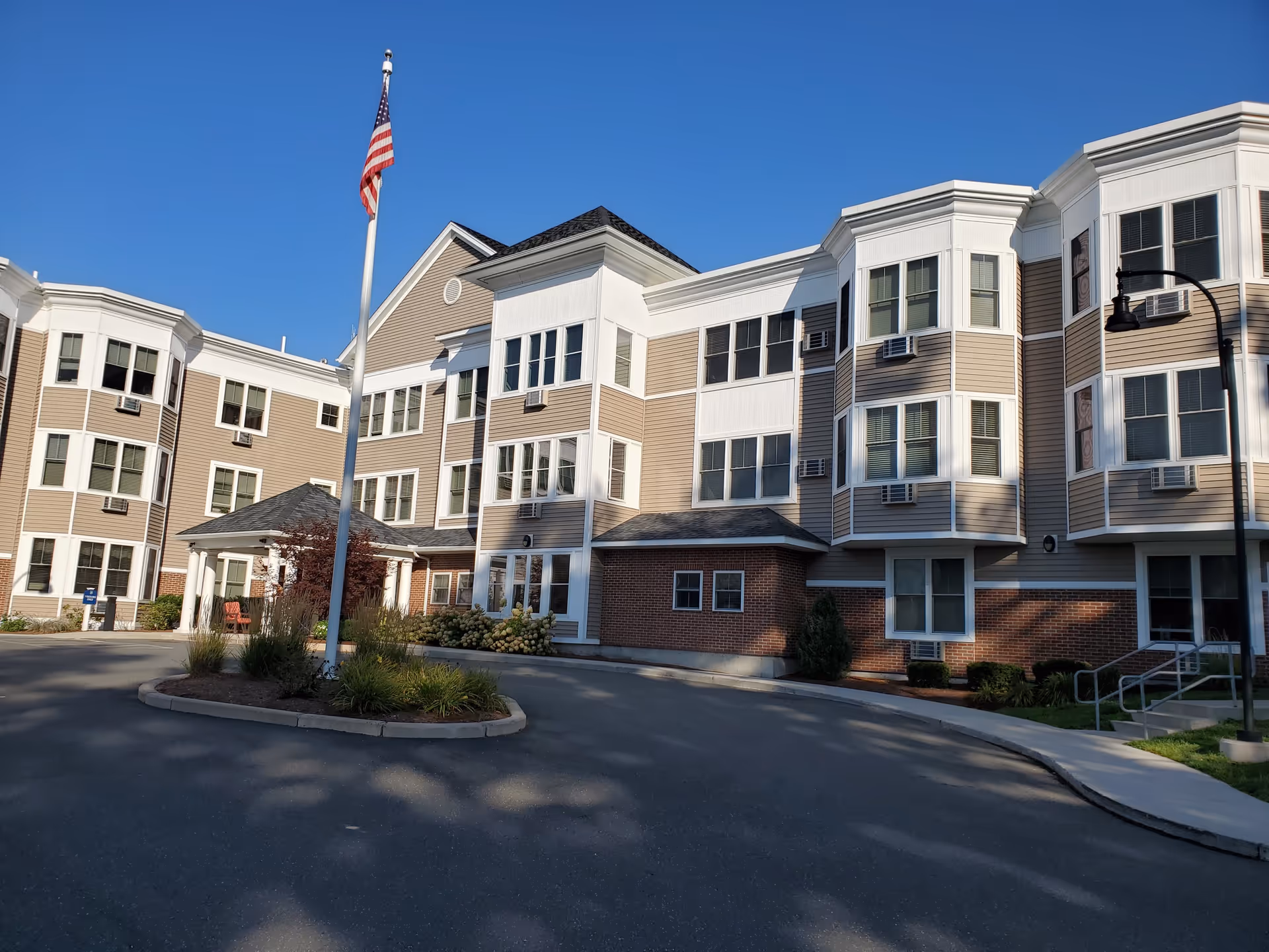 Exterior view of a multi-story senior living facility building with beige siding, white trim, and multiple windows. There is a flagpole with an American flag in front of the building, surrounded by landscaping and a circular driveway under a clear blue sky.