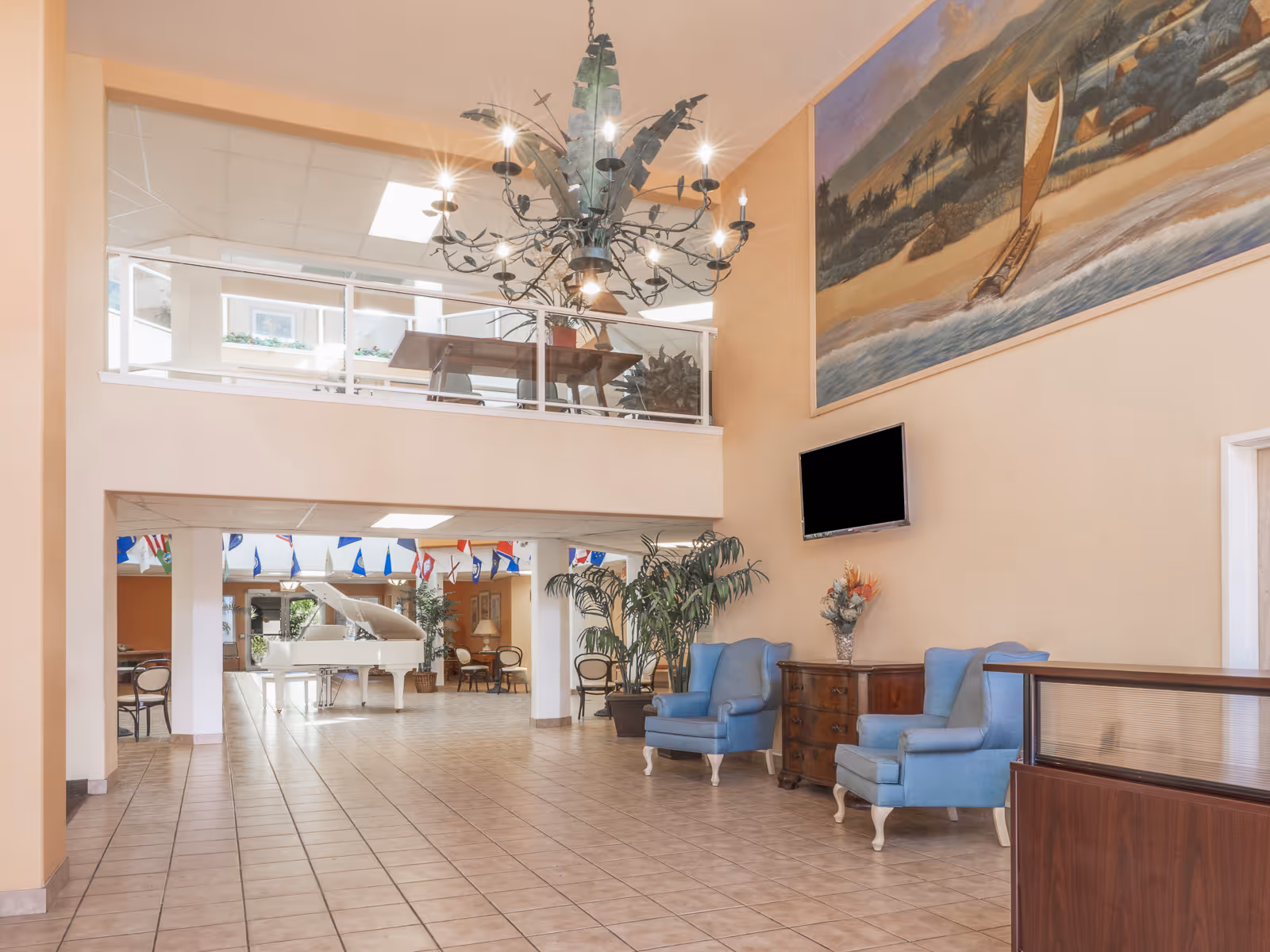 Spacious senior living facility common area with tiled floor, two blue armchairs, a wooden chest with a flower vase, a large wall-mounted TV, and a large mural painting above. In the background, a white grand piano is visible under a mezzanine with glass railing and a decorative chandelier hanging from the ceiling.