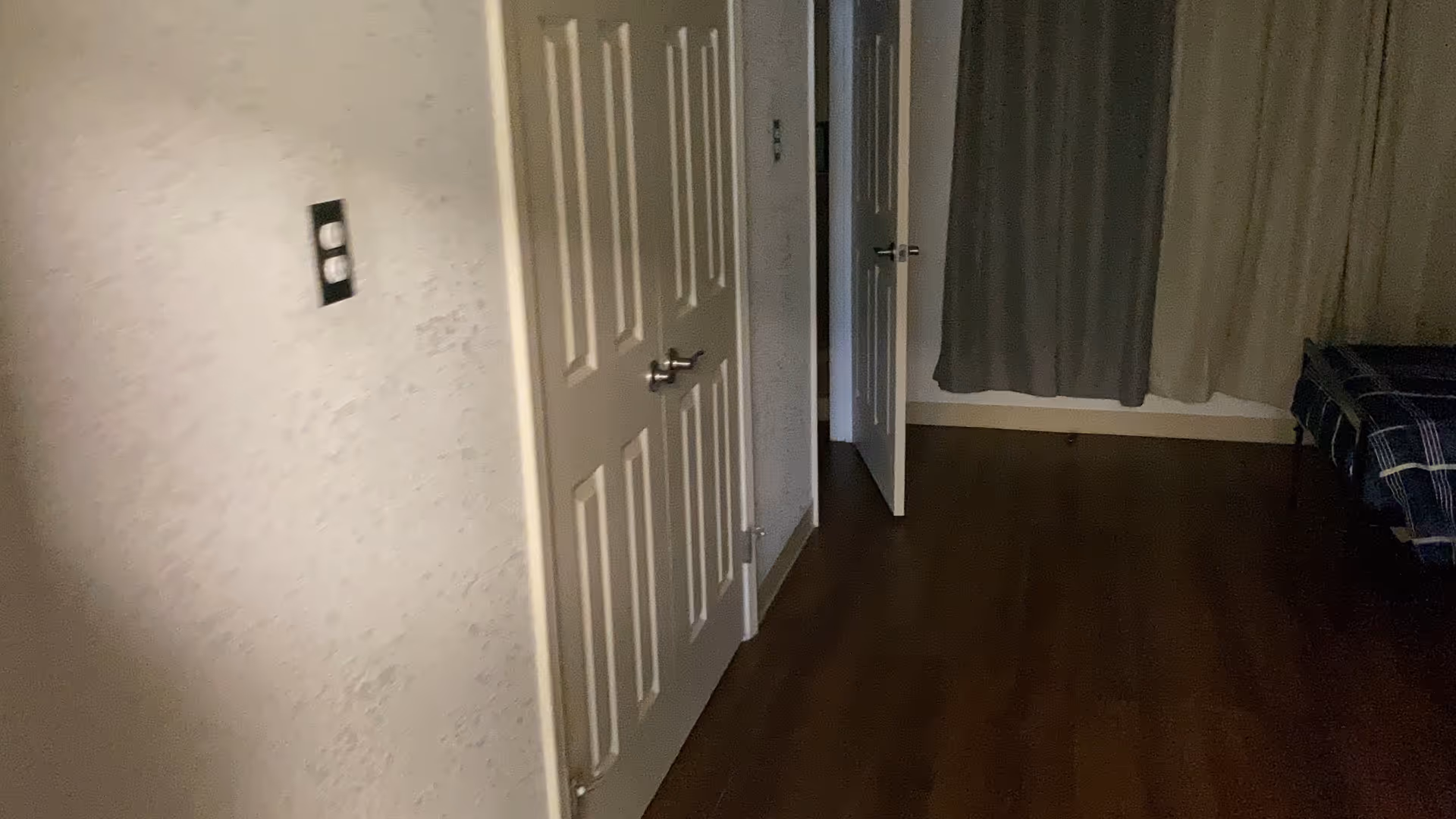Dimly lit bedroom interior showing closet doors, an open doorway, curtains, hardwood floor, and part of a bed.