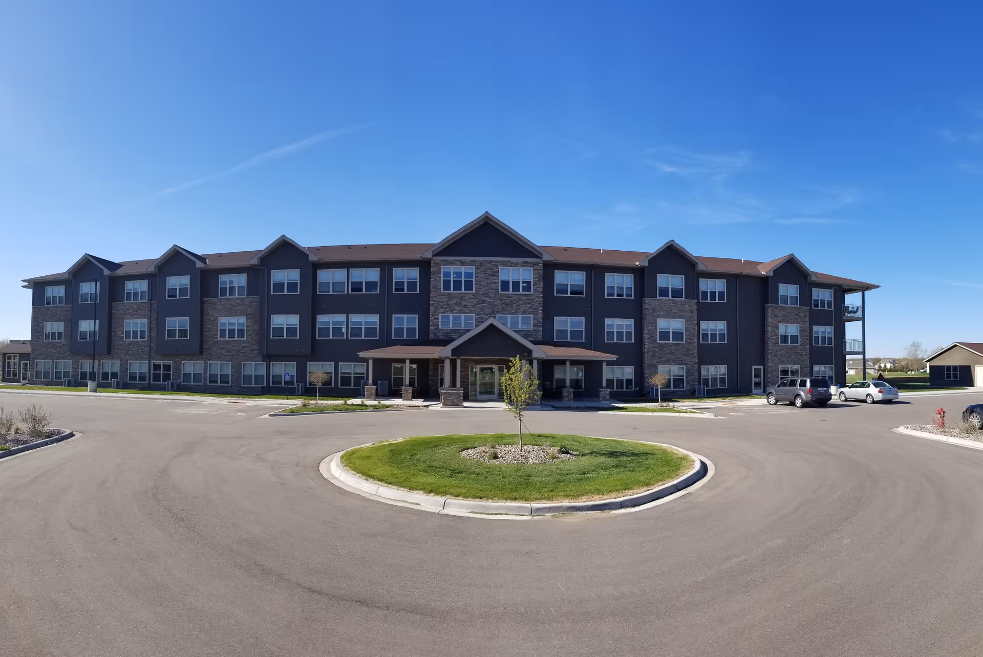 Front exterior view of a three-story senior living facility building with a circular driveway and a small landscaped island with a young tree in the center. The building has multiple windows and a covered entrance under a peaked roof. Several cars are parked near the building under a clear blue sky.