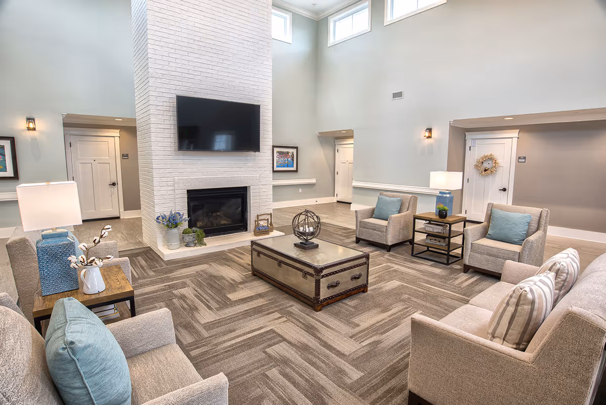 A spacious and well-lit living room area with beige armchairs and a sofa adorned with light blue and striped pillows. A large white brick fireplace with a mounted flat-screen TV is the focal point. The room features a patterned carpet, side tables with lamps and decorative items, and high windows near the ceiling allowing natural light in.