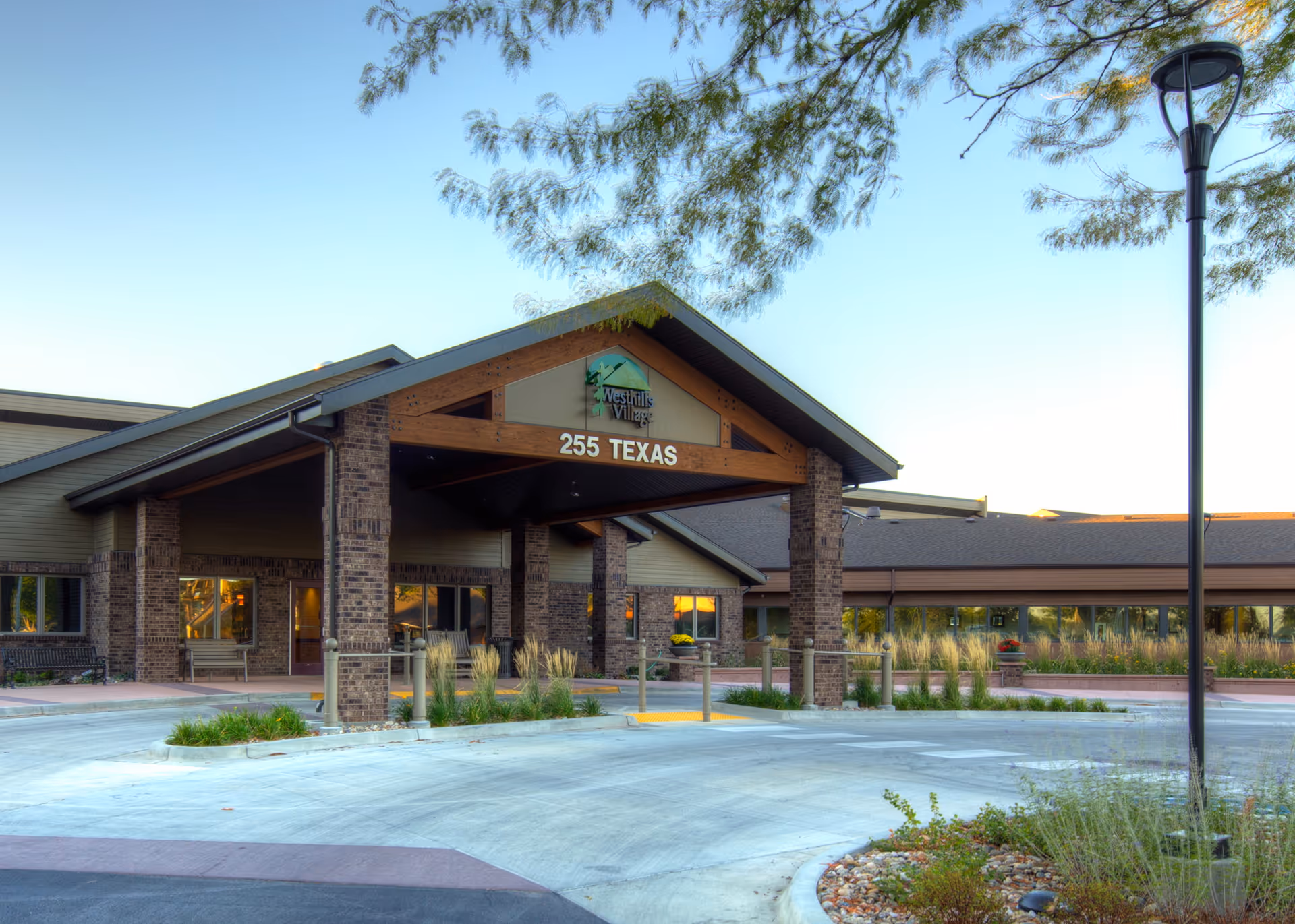 Exterior front entrance of Westhills Village facility with a covered drop-off area supported by brick pillars, landscaping with plants and grasses, and a street lamp on the right side.