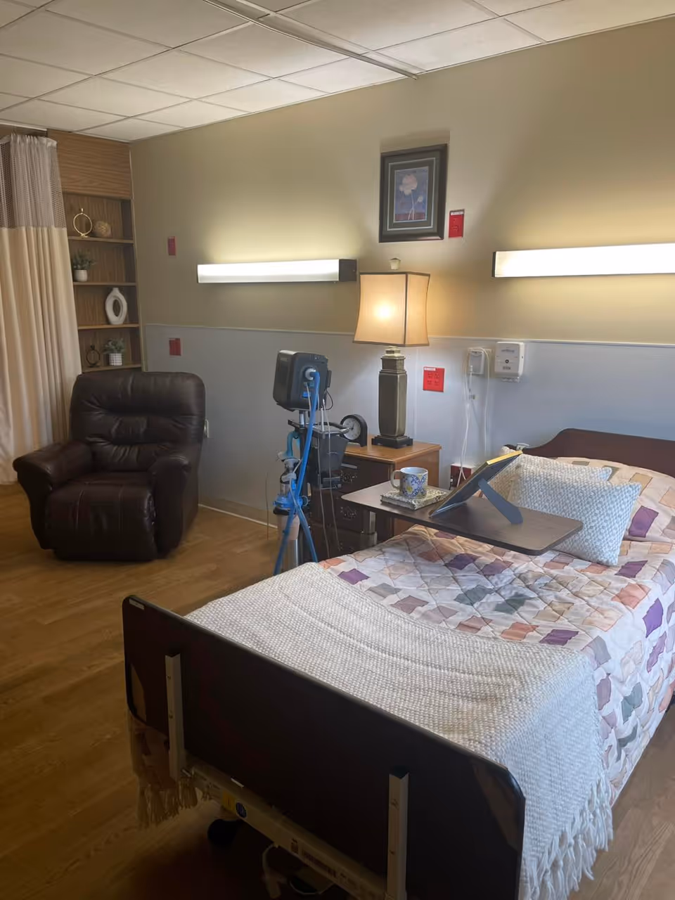 A senior care facility room with a hospital bed covered in a colorful quilt and a white blanket. There is a pillow and a small overbed table with a cup and a tablet stand on it. Next to the bed is a wooden nightstand with a lamp and medical equipment. A brown recliner chair is positioned near a curtain and a wooden shelving unit with decorative items. The room has wood flooring and soft lighting from wall-mounted fixtures.