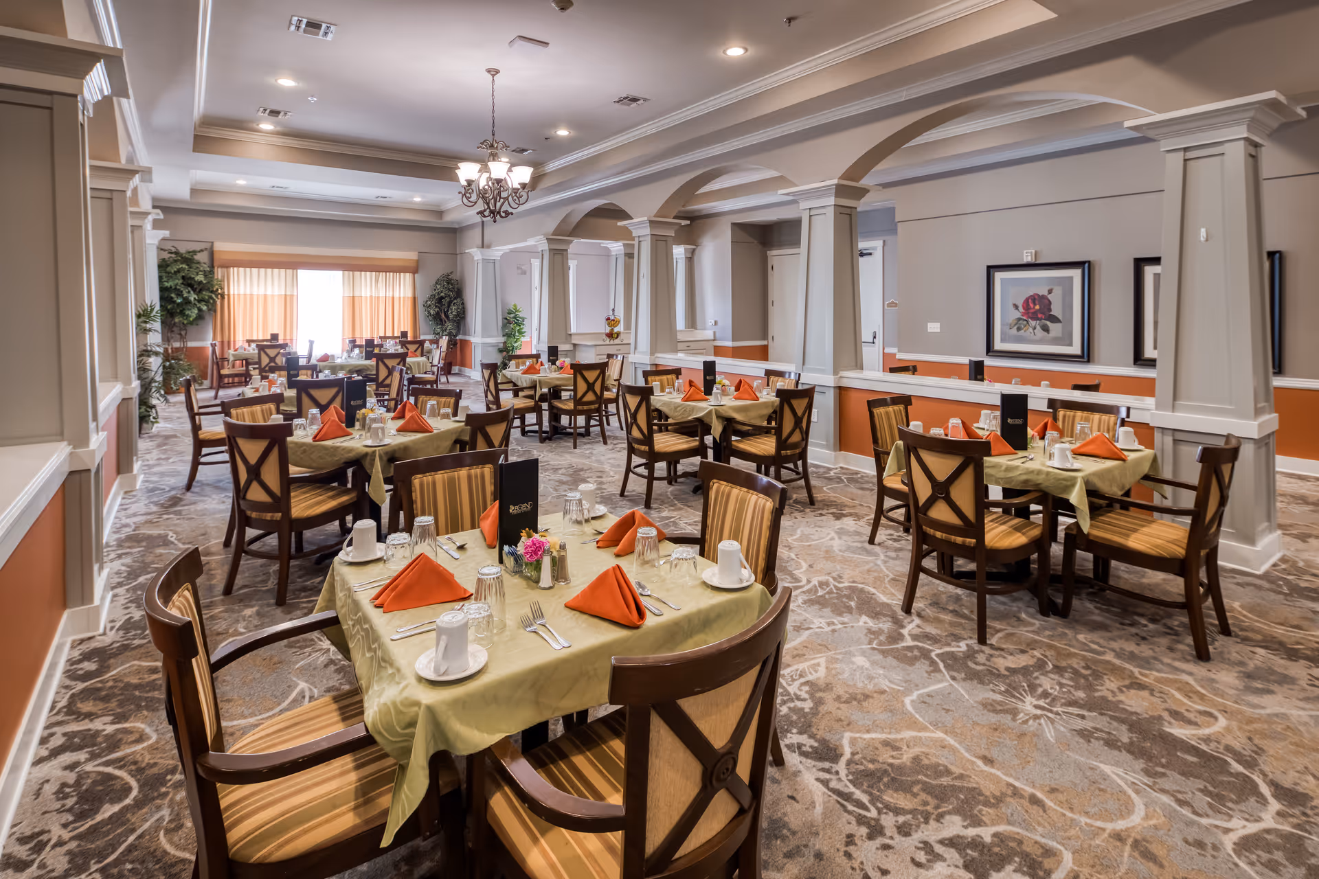 A spacious dining room with multiple tables set for a meal. Each table has a green tablecloth, orange folded napkins, white cups, glasses, and silverware. The room features beige walls with white trim, decorative columns, a chandelier, framed artwork, and large windows with curtains letting in natural light.