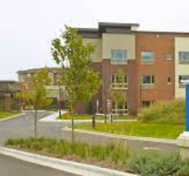 Exterior view of a modern senior living facility building with a driveway, landscaped greenery, and young trees in front.