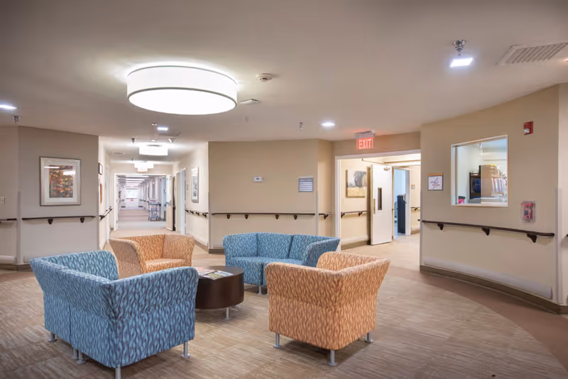 A well-lit common area in a senior living facility with four upholstered chairs arranged around a small round table. The walls are beige with handrails, and there are framed pictures hanging. A hallway extends into the distance with multiple doors and ceiling lights. An exit sign is visible above a doorway on the right.