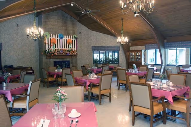 Dining room with multiple tables covered in pink tablecloths, each set with glassware, plates, and silverware. The room features wooden ceiling beams, chandeliers, a fireplace with decorative flowers and colorful fans above it, and windows letting in natural light.