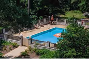 Outdoor rectangular swimming pool with lounge chairs, umbrellas and a fenced patio surrounded by trees and landscaping.