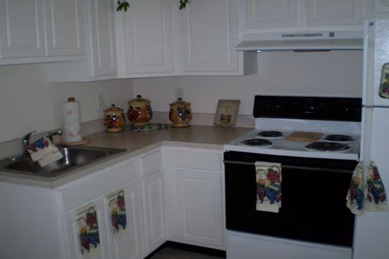 A small kitchen corner with white cabinets and countertops. There is a stainless steel sink with a faucet on the left, a white electric stove with four burners in the center, and a white refrigerator on the right. The countertop has three decorative canisters and a framed picture. Several kitchen towels with grape designs hang on the cabinet doors and stove handle.