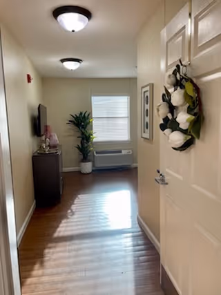 View down a hallway in an assisted living facility with wooden flooring, beige walls, and two ceiling lights. On the right side, a door is decorated with a wreath of white flowers. At the end of the hallway, there is a window with blinds, a potted plant, a wall-mounted air conditioning unit, a framed picture on the wall, and a small cabinet with a television on top.
