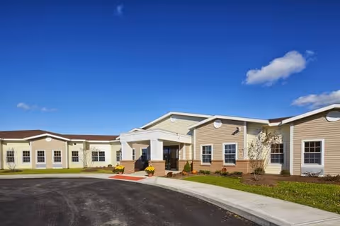 Exterior view of St. Francis Commons Assisted Living Residence showing a single-story building with beige and light yellow siding, a covered entrance, and a paved driveway under a clear blue sky with a few clouds.