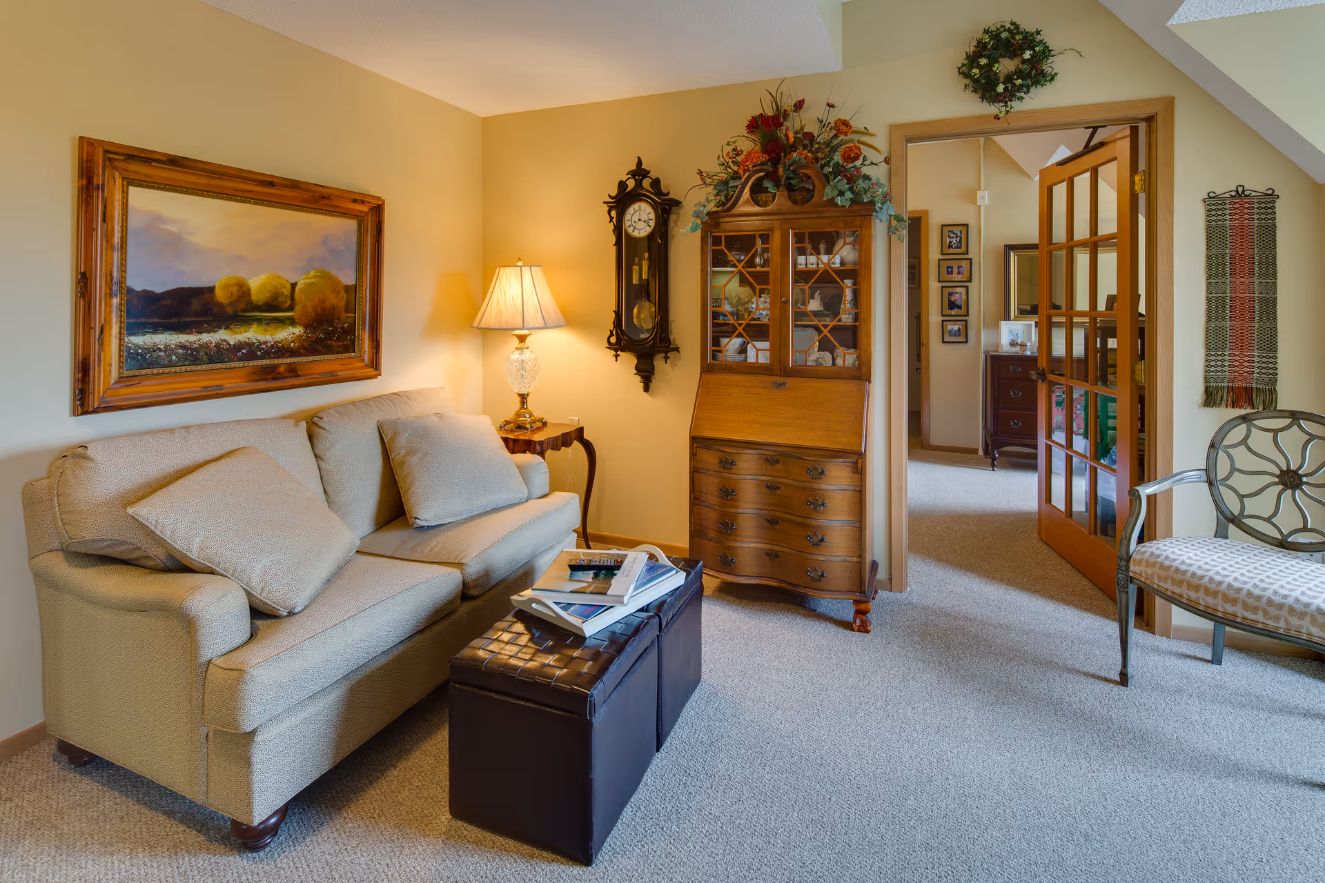 A cozy living room with a beige sofa adorned with cushions, a wooden side table with a lamp, a wall-mounted antique clock, and a wooden cabinet with glass doors displaying decorative items. A framed landscape painting hangs above the sofa, and a cushioned chair with a metal frame is positioned near a doorway with glass-paneled double doors leading to another room. The room has beige walls and carpeted flooring.