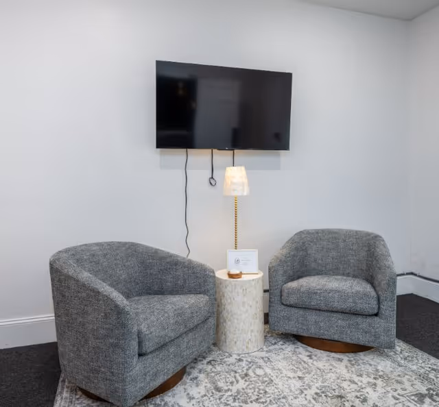 A small seating area with two gray upholstered armchairs positioned on a patterned rug. Between the chairs is a small round side table with a lamp and a framed item on it. A flat-screen TV is mounted on the white wall above the table, with visible cords hanging down.