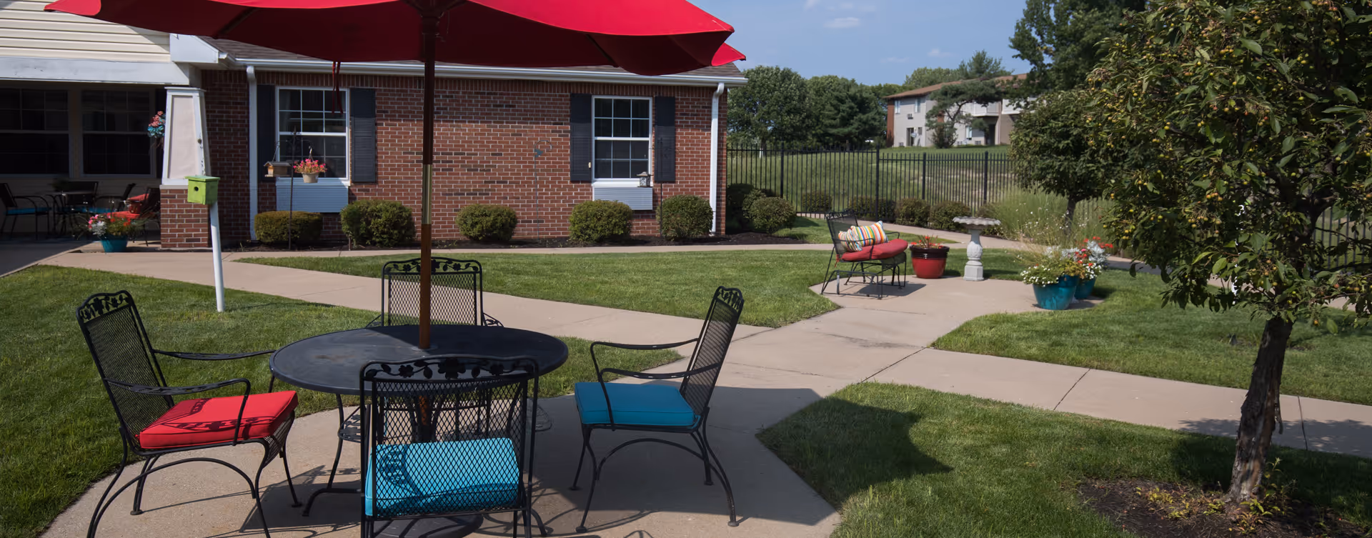 Sunlit outdoor courtyard with a round table and umbrella, cushioned metal chairs, walkways, potted plants and a brick building in the background.