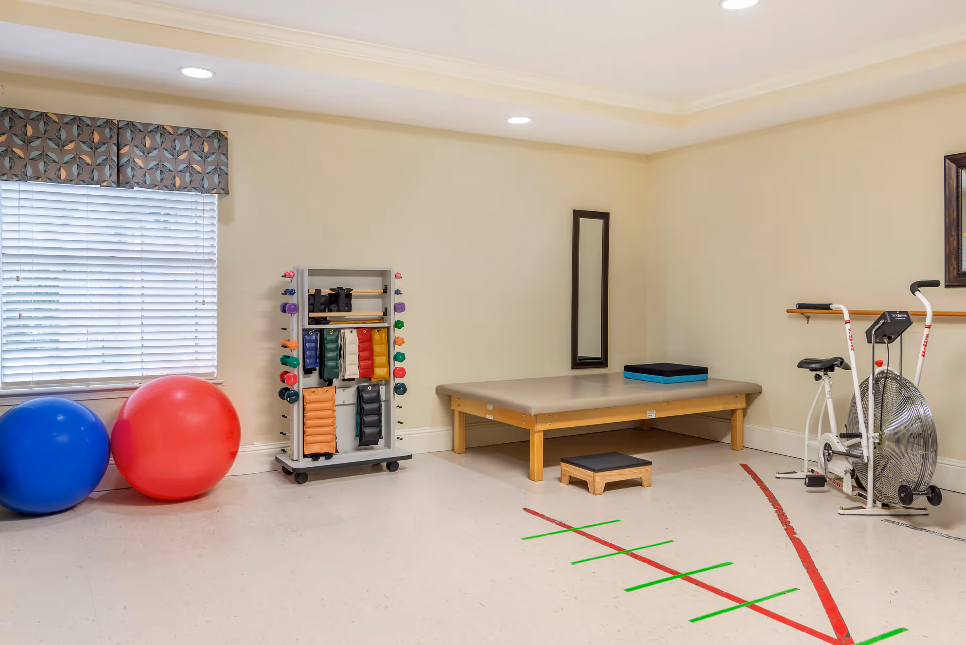 A physical therapy or exercise room with a large blue and red exercise ball, a rack holding various small weights and exercise equipment, a padded therapy table with a step stool, a stationary exercise bike, and a wall-mounted mirror. The room has beige walls, a window with blinds and a patterned valance, and a floor marked with red and green tape for exercise guidance.