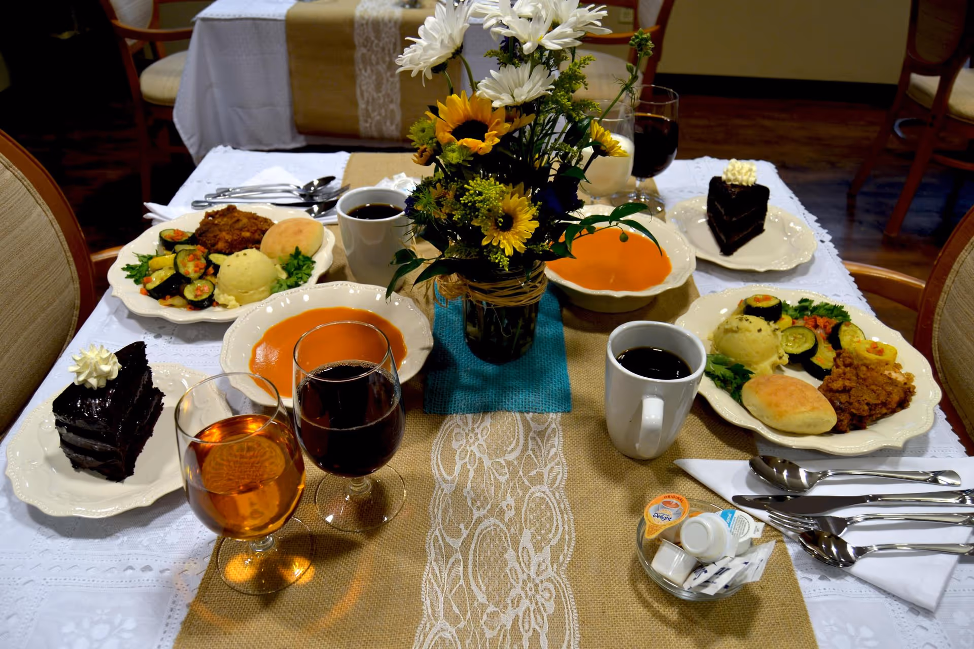 A dining table set with plates of food including fried chicken, mashed potatoes, vegetables, rolls, bowls of soup, slices of chocolate cake with whipped cream, glasses of iced tea and red wine, cups of coffee, and a small vase with a bouquet of yellow and white flowers in the center.