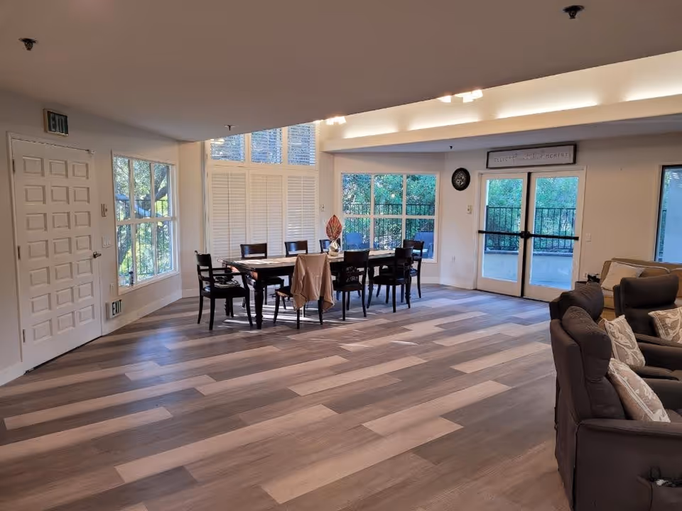 A spacious interior room with a wooden dining table surrounded by eight chairs, a large window with white shutters, glass doors leading outside, and a seating area with cushioned chairs and sofas. The floor has a wood pattern, and the room is well-lit with natural light coming through the windows.
