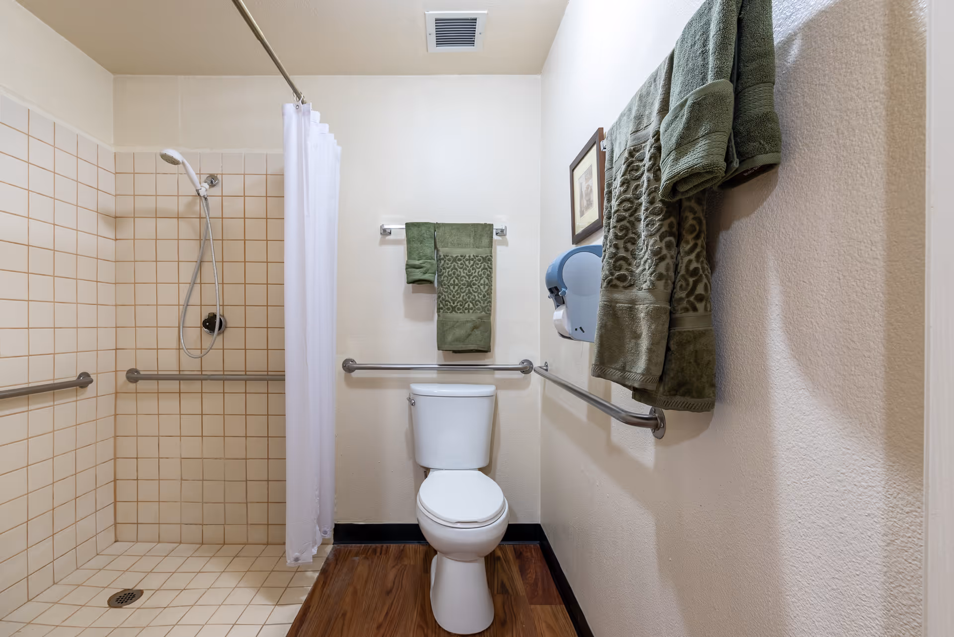 A clean and accessible bathroom featuring a tiled walk-in shower with a handheld showerhead and grab bars, a white toilet with grab bars on both sides, green towels hanging on the wall, a wall-mounted paper towel dispenser, and a framed picture.