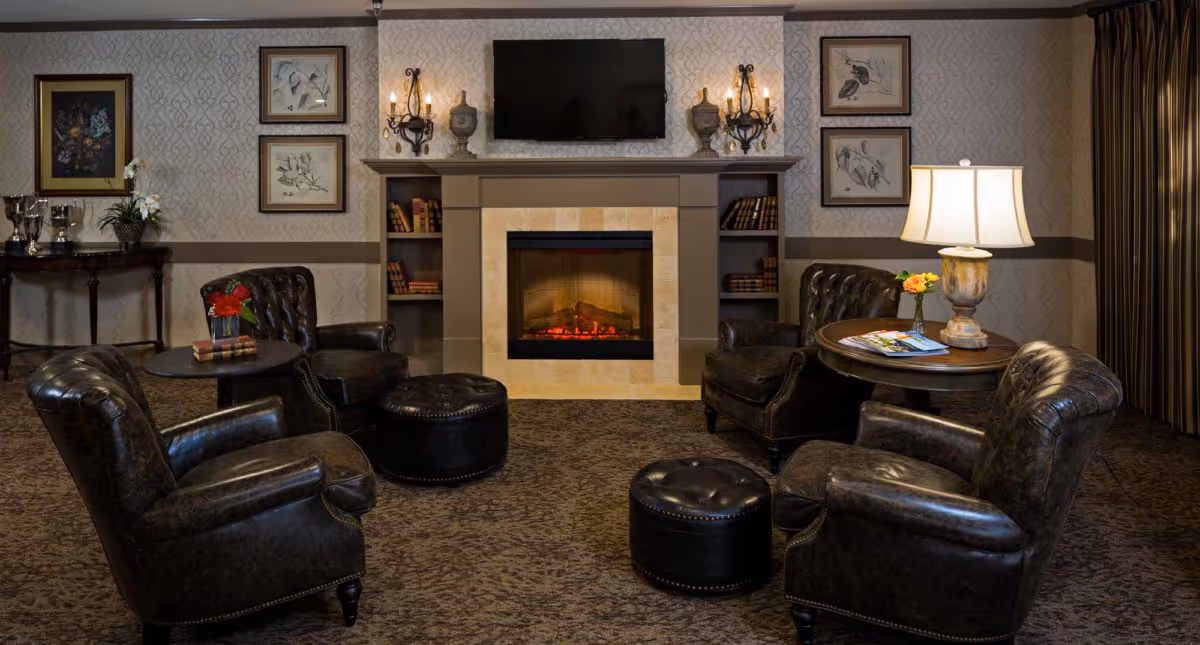 A cozy living room area with four dark leather armchairs arranged around two small round tables and two matching ottomans. The room features a fireplace with a TV mounted above it, built-in shelves with books, wall sconces, framed artwork, a table lamp, and decorative items on a side table against the wall.