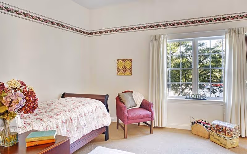 A bright bedroom with a single bed covered in a floral quilt, a wooden nightstand with books and a vase of flowers, a pink upholstered armchair with a pillow and blanket, a large window with cream curtains letting in natural light, and decorative baskets and boxes on the floor near the window.