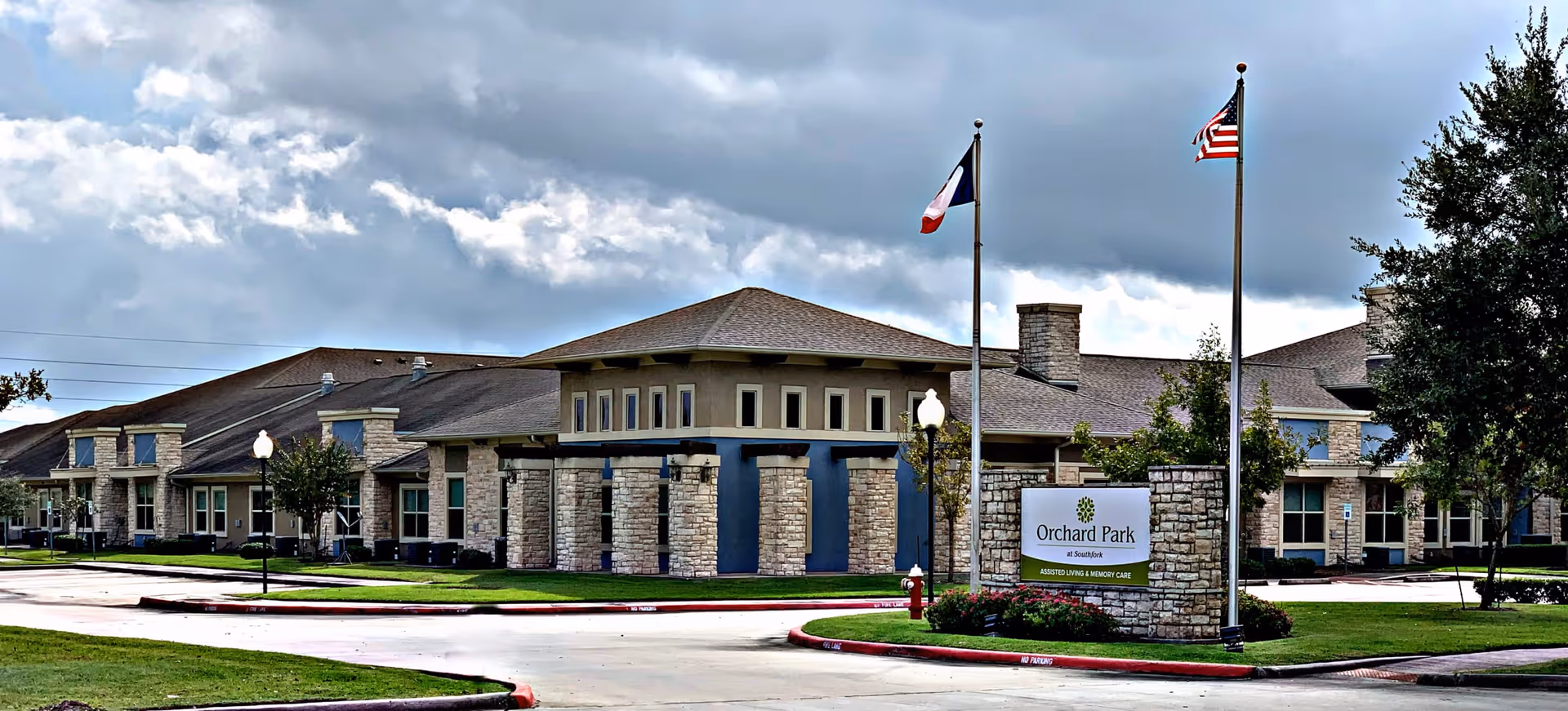 Exterior view of Orchard Park at Southfork Assisted & Senior Living facility showing a single-story building with stone and beige walls, two flagpoles with the Texas state flag and the American flag, a sign with the facility name, and a cloudy sky above.
