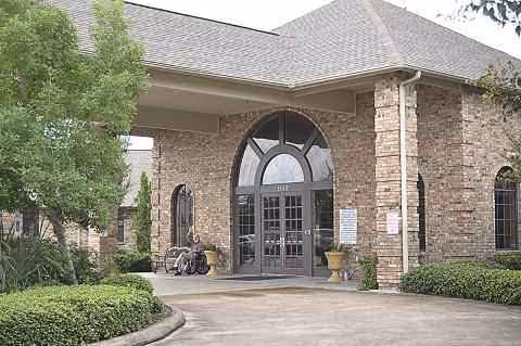 Brick facility entrance with arched glass doors, columns, and potted plants along a curved walkway.