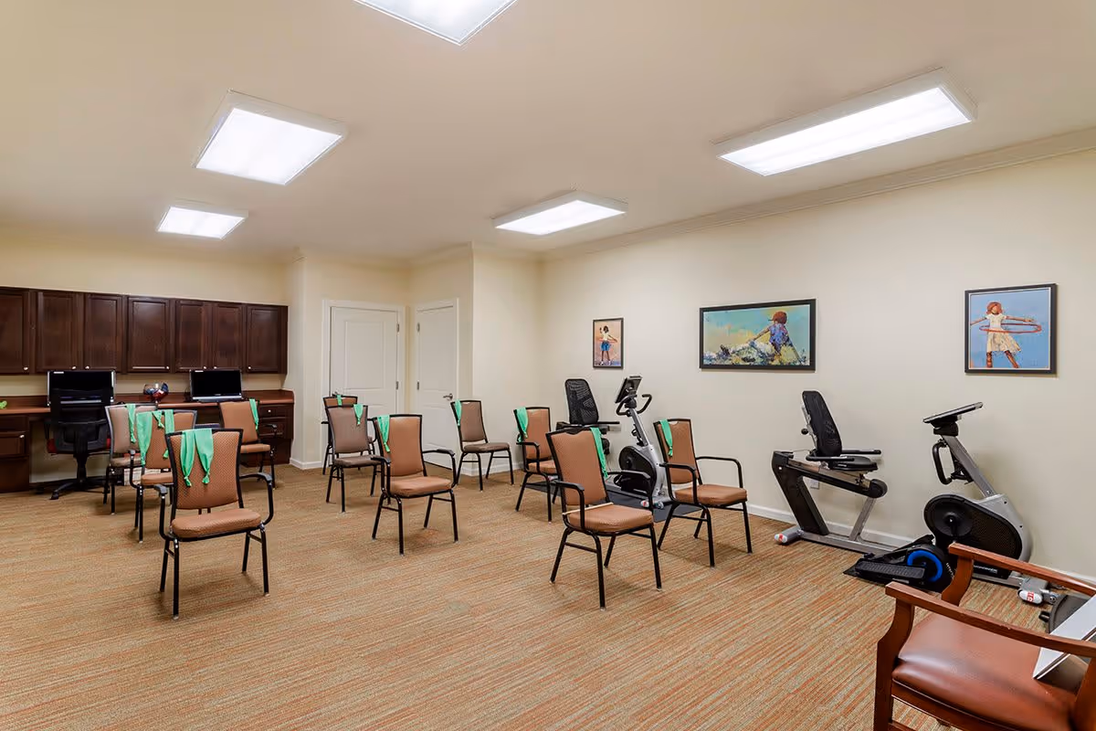 A spacious room with several chairs arranged in rows, each chair draped with a green towel. The room has exercise equipment including stationary bikes along one wall, and three framed pictures of children playing on the wall above the equipment. There are built-in desks with computers and office chairs along the back wall. The room is well-lit with ceiling lights and has beige walls and carpeted flooring.