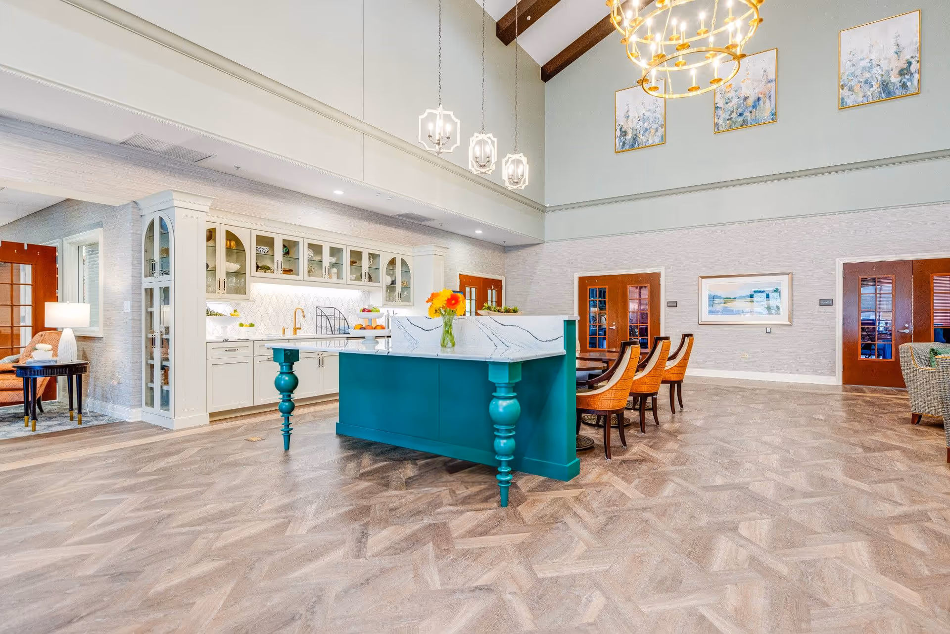 A bright and spacious interior room featuring a teal kitchen island with a white marble countertop and a vase of flowers on top. Behind the island is a white cabinet with glass doors and a gold faucet. To the right, there is a dining area with a table and orange cushioned chairs. The room has high ceilings with exposed wooden beams and modern hanging light fixtures. The walls are decorated with framed artwork, and there are wooden doors with glass panels leading to other rooms.