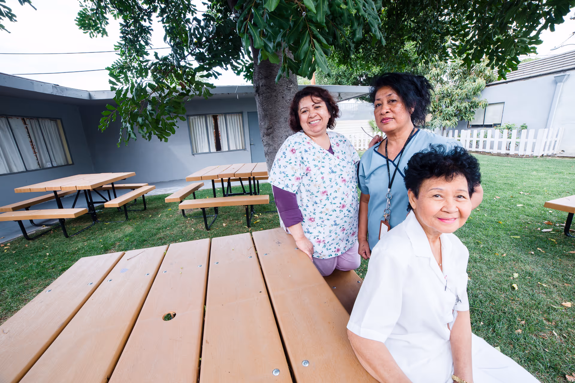 Three women, two standing and one sitting, smiling in an outdoor garden area with picnic tables and a tree, surrounded by a building with windows and a white picket fence.