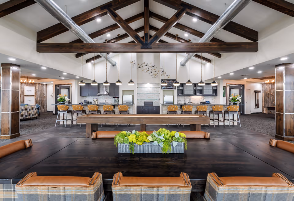 Spacious communal dining and kitchen area with a long wooden table, bar seating, exposed ceiling beams, and a floral centerpiece.