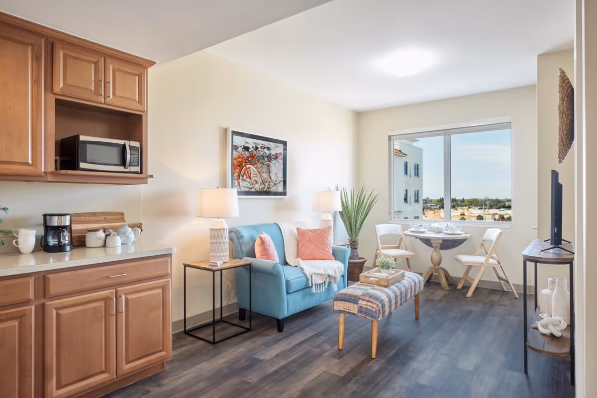 A bright and cozy living area in a senior living facility featuring a small blue loveseat with pink pillows and a white throw, two table lamps on either side, a small wooden side table, a patterned bench, a round dining table with two chairs near a large window, and a kitchen counter with wooden cabinets, a microwave, and a coffee maker.