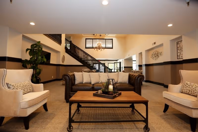 A spacious and well-lit living room area featuring a dark brown leather sofa with multiple light-colored cushions, flanked by two beige armchairs with patterned pillows. A wooden coffee table with a decorative tray and candle sits in front of the sofa. In the background, there is a staircase with dark wooden railings, a chandelier hanging from the ceiling, and large windows allowing natural light to enter.